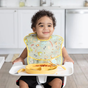 A smiling curly-haired toddler sits in a high chair, wearing a Bumkins SuperBib®: Camp Gear, with food on the tray.