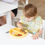 A toddler in a Bumkins SuperBib®: Camp Gear uses a fork with a yellow plate featuring a camping print.