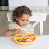 Child with curly hair beams while eating from an orange plate, seated on a white chair in their Bumkins SuperBib®: Camp Gear.