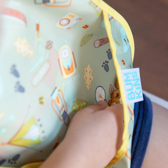 A childs hand fills the SuperBib®: Camp Gear pocket with snacks. Bumkins tag gleams on its waterproof fabric.