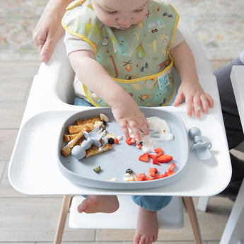 Baby in high chair playing with strawberries and toast, wearing a Bumkins SuperBib®: Camp Gear with a playful camping print.