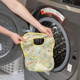 Someone is placing a Bumkins SuperBib®: Camp Gear into a front-loading washing machine.