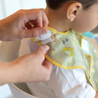 Hands fasten a Bumkins SuperBib®: Camp Gear around a smiling baby seated in a high chair, enjoying the waterproof fabric.