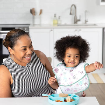 A smiling adult and child in the kitchen; the child points, sitting with a blue plate wearing a Bumkins SuperBib®: Floral.