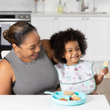 A woman smiles with a child in a Bumkins SuperBib®: Floral, holding a snack by the blue plate.