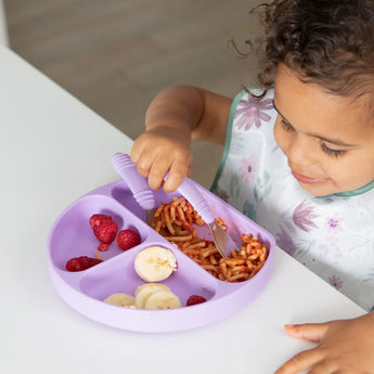 A young child in a colorful Bumkins SuperBib®: Floral enjoys raspberries, spaghetti, and bananas from a purple divided plate at the table.