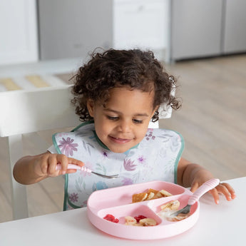 A curly-haired child eats from a pink divided plate, wearing a Bumkins SuperBib®: Floral, sitting at a white table.