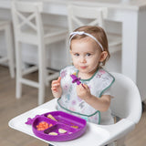 A toddler in a high chair, wearing the Bumkins SuperBib®: Floral, holds a purple spoon in front of a divided plate.