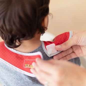 An adult fastens a red and gray SuperBib® from Bumkins Super Mario™ Power-Up pack, showcasing its easy-wipe fabric.