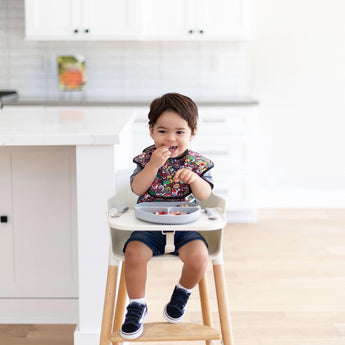 A toddler in a Bumkins SuperBib® 3 Pack: Super Mario™ Power-Up sits on a high chair, enjoying snacks with easy-wipe fabric nearby.