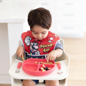 A toddler in a high chair enjoys food from a red plate, wearing an easy-wipe Bumkins Super Mario™ Power-Up SuperBib.