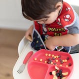 Toddler in a high chair wearing a Bumkins SuperBib® with Super Mario™, picks snacks from a pink divided plate with leftovers.