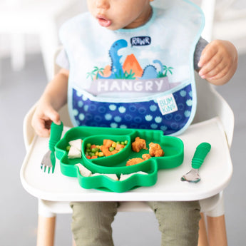 Toddler in a high chair with a blue dinosaur bib, happily using Bumkins Spoon + Fork: Jade and a green dinosaur plate.