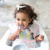 A toddler in a high chair, wearing a Bumkins SuperBib: Magical World, smiles holding a spoon with a bowl of food in front.