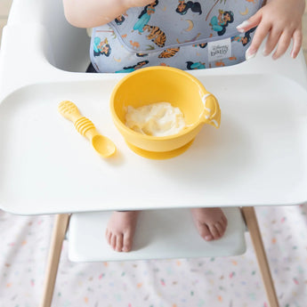 A baby in a high chair with a yellow bowl and spoon wears a Bumkins SuperBib® from the Magical World, Ariel, and Jasmine pack.