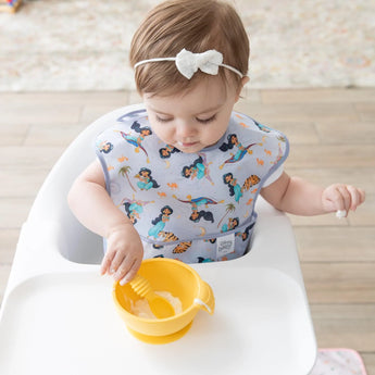 Toddler in high chair using Bumkins SuperBib® from the 3-Pack (Magical World, Ariel, Jasmine), reaching into yellow bowl with fork.