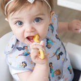 A blue-eyed toddler enjoys a meal, wearing a Bumkins SuperBib®: Ariel design, with a yellow spoon.