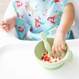 A child in a Bumkins SuperBib® enjoys yogurt with strawberries from a green bowl while sitting in a high chair.