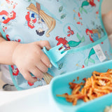 A child holds a teal fork and wears a Bumkins SuperBib® featuring Ariel, with noodles on the table.