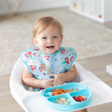 A toddler in a Bumkins SuperBib® enjoys a meal from a blue divided plate in a bright, minimalistic room.