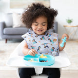 A curly-haired toddler in a high chair dons a Bumkins SuperBib® featuring Ariel while using a spoon with a divided plate.