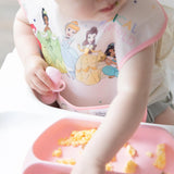 A baby in a Bumkins SuperBib® (Ariel design) with a pink pacifier reaches for scrambled eggs on a waterproof fabric tray.