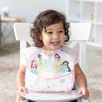 Toddler smiling happily, seated with a Bumkins SuperBib® from the 3-pack featuring Ariel, Jasmine, and Magical World designs.