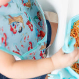 A child in a Bumkins SuperBib® featuring Disneys Ariel and Jasmine, joyfully eating spaghetti from a blue tray in their high chair.