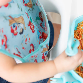 A child in a Bumkins SuperBib® featuring Disneys Ariel and Jasmine, joyfully eating spaghetti from a blue tray in their high chair.