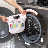 A person holds a pink Disney princess-themed SuperBib from the Bumkins SuperBib® 3-Pack featuring Magical World in front of an open washing machine. The bib displays animated princesses and reads, Its a Magical World, as the machine door stands ready.