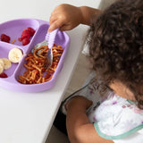 A child enjoys spaghetti, bananas, and raspberries on a purple plate, wearing a Bumkins SuperBib®: Floral.