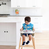 A toddler in a high chair wears a Mischief Managed bib, eating fruit from a Bumkins Silicone Grip Dish in gray.