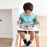 A toddler enjoys fruit and cereal from a Bumkins Silicone Grip Dish in gray with a suction base on a light wooden floor.