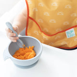 A baby in a yellow Desert Boho SuperBib® by Bumkins holds a spoon over orange mash on a white highchair tray.