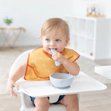 A blonde baby sits in a high chair wearing a Bumkins SuperBib® (Desert Boho), chewing a spoon next to a bowl in a bright room.