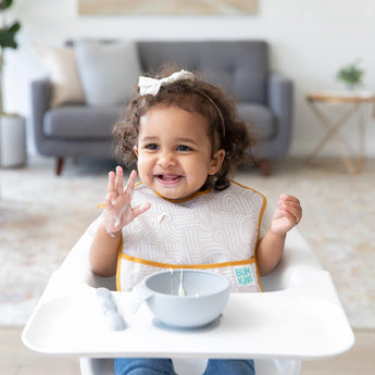 Smiling toddler in a high chair, wearing a Bumkins SuperBib® from the Desert Boho Collection, plays with food in a cozy setting.