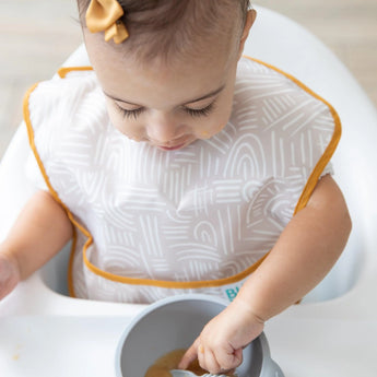 A toddler in a high chair uses a Bumkins SuperBib and spoon to eat from a bowl, with a matching bow from the Desert Boho collection.