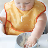 Baby in high chair wearing Bumkins SuperBib® from the Desert Boho 3 Pack, playfully smeared with creamy delight on mouth and hands.