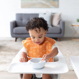 A toddler in a Bumkins SuperBib® 3 Pack: Desert Boho eats happily in a high chair, with the cozy living room collection glowing behind.