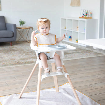 A baby in a high chair, wearing a Bumkins SuperBib® Desert Boho, holds utensils in a bright, modern living room.