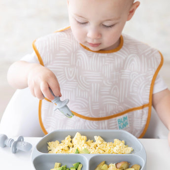 A baby enjoys scrambled eggs with a gray fork on a plate, using the Bumkins SuperBib® 3 Pack: Desert Boho, avocado, and potatoes.