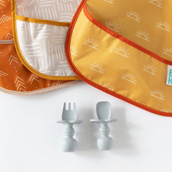 Desert Boho SuperBib® 3 Pack by Bumkins with sun patterns and gray toddler utensils on a white surface.