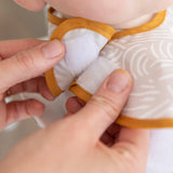 Close-up of hands fastening a Bumkins SuperBib from the Desert Boho collection—white, waterproof with yellow trim and a subtle pattern.