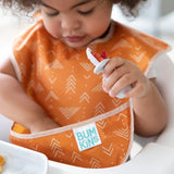 A toddler uses a fork while wearing Bumkins waterproof SuperBib® from the Desert Boho collection, featuring an orange pattern.