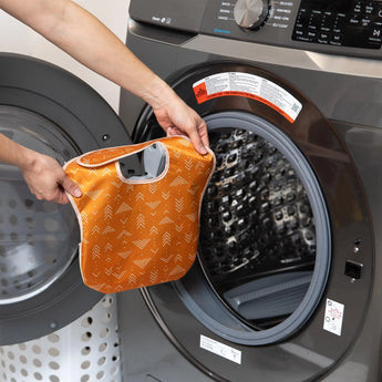 Someone places a Bumkins SuperBib® Desert Boho wet bag into an open front-loading washing machine.