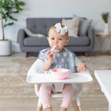 A toddler with a white bow sits in a high chair using a spoon with her Bumkins SuperBib®, eating from a pink bowl, gray couch behind.