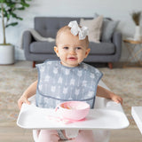 A baby with a bow sits in a high chair, wearing Bumkins waterproof SuperBib: Arrow, with a bowl and spoon on the tray.