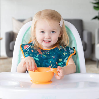 A toddler sits in a high chair, wearing a Bumkins SuperBib® from the All Together Now set, and touching an orange floral bowl.