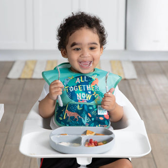 A smiling toddler in a high chair wears a Bumkins SuperBib® from the All Together Now pack, holding cutlery with a meal tray ahead.