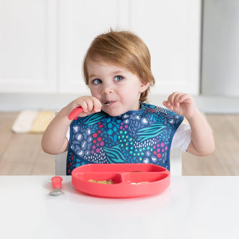 A brown-haired child wears a Bumkins SuperBib®, holding a spoon, and eats from a red divided plate at the table.